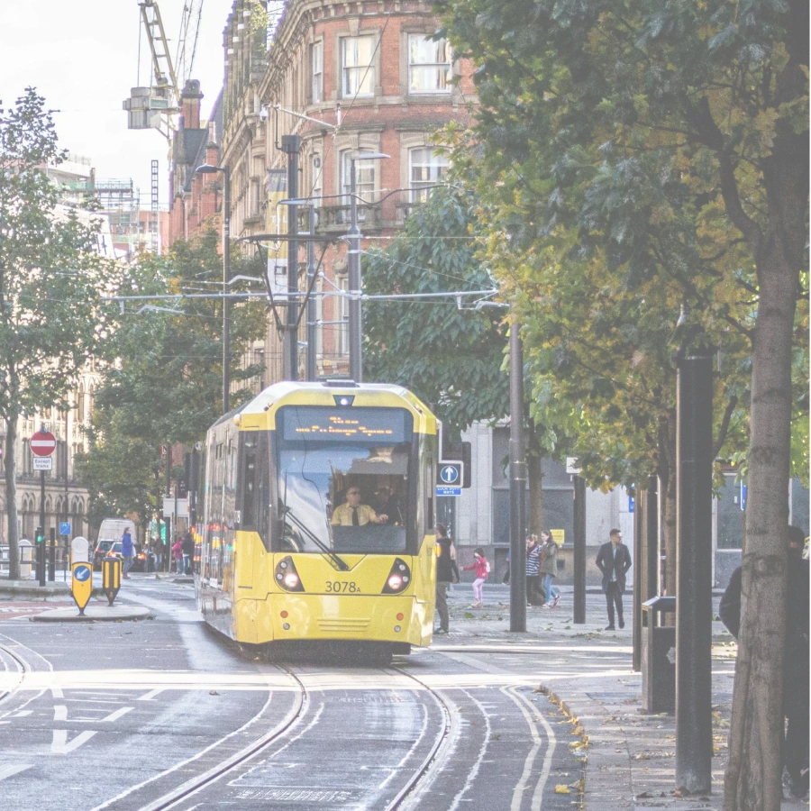 A yellow Metrolink tram on a tree-lined Manchester street.